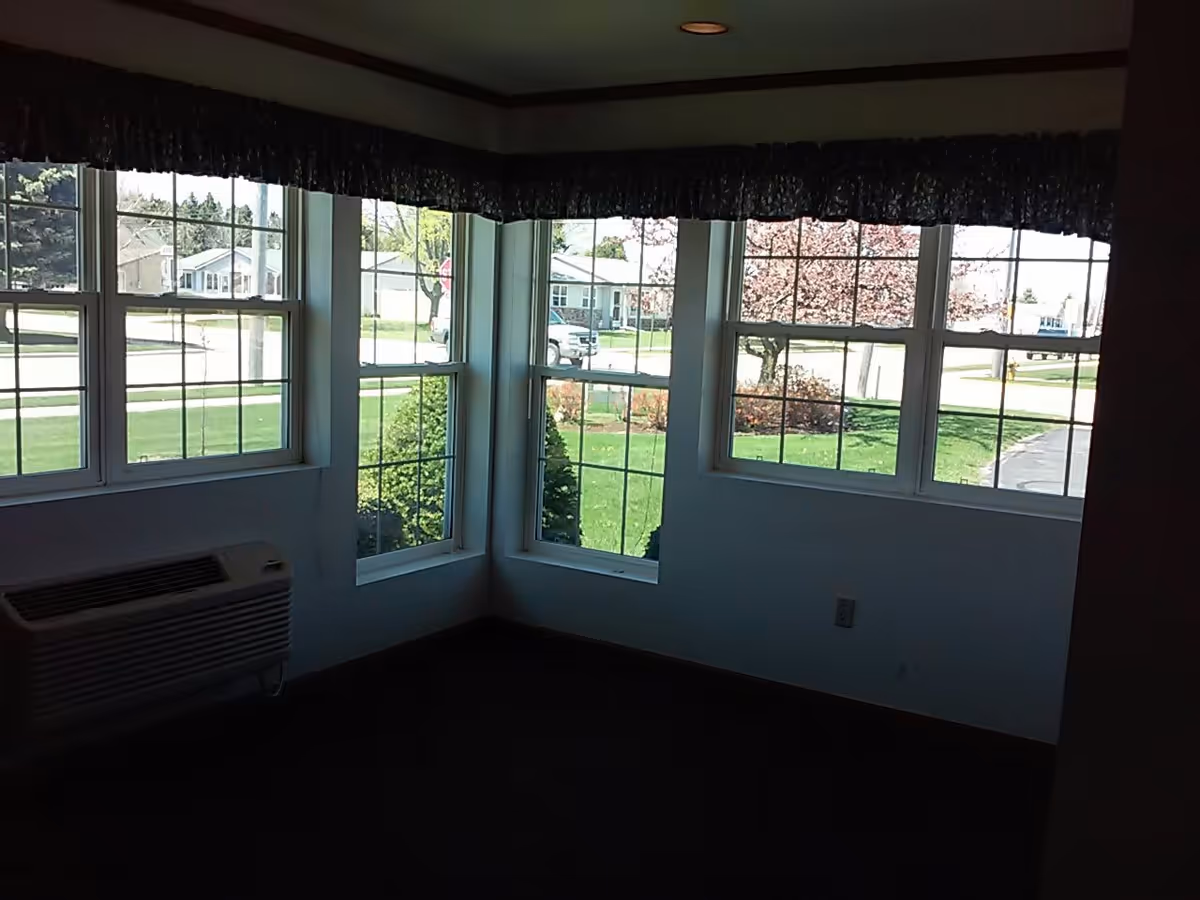 Interior corner of a room with three large windows showing a green lawn, trees, and houses outside. The room has dark carpet, white walls, a wall-mounted air conditioning unit, and a dark valance above the windows.