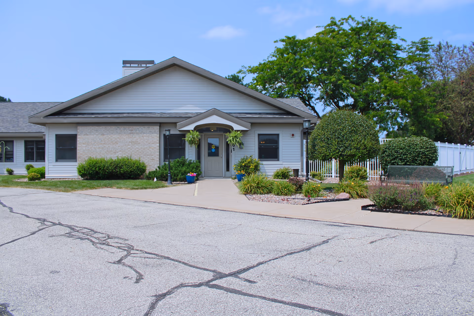 Front exterior view of a single-story building with a peaked roof, a central entrance door flanked by windows, hanging plants, and landscaped bushes and flowers along the walkway leading to the entrance. The sky is clear and blue.