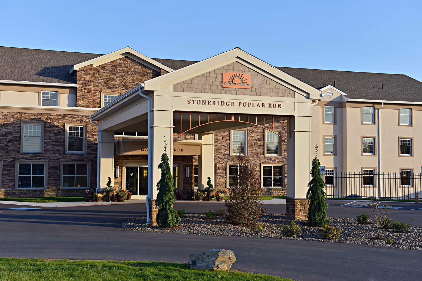 Exterior view of the entrance to Stoneridge Poplar Run, a retirement living community. The building features a covered drop-off area with stone and beige siding, multiple windows, and landscaped greenery around the driveway.