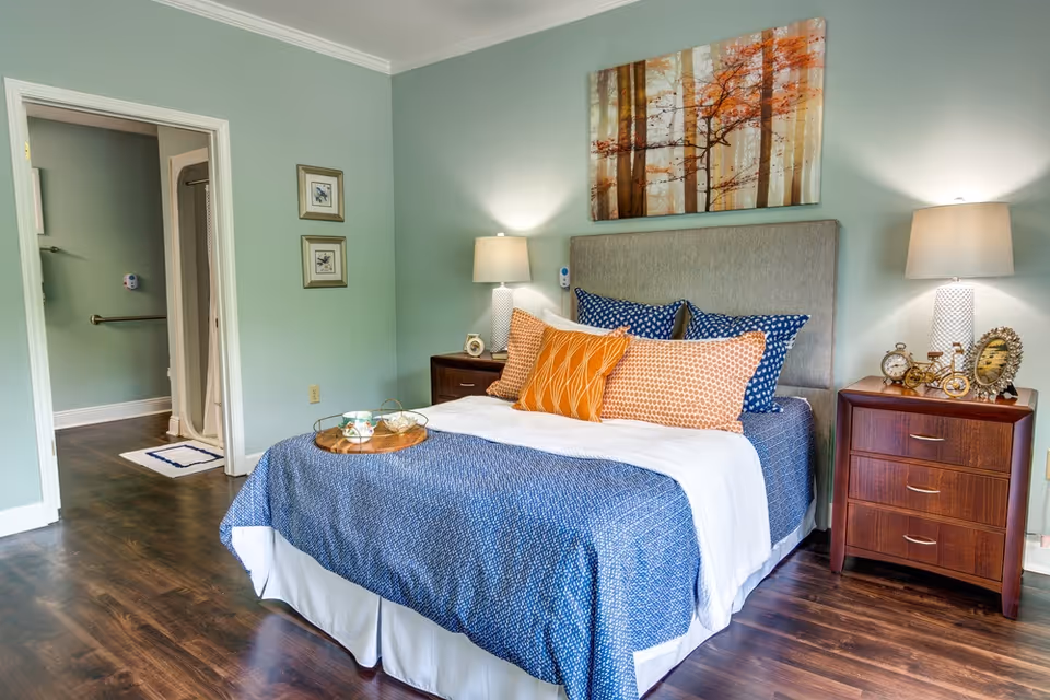 A neatly made bed with a blue bedspread, white blanket, and multiple decorative pillows in orange and blue patterns. The bed has a gray upholstered headboard and is flanked by two wooden nightstands, each with a lamp. On the right nightstand, there are decorative items including a small clock and a framed photo. Above the bed hangs a painting of trees with autumn-colored leaves. The room has light green walls and dark wood flooring. An open doorway on the left leads to a bathroom with a shower and grab bars.