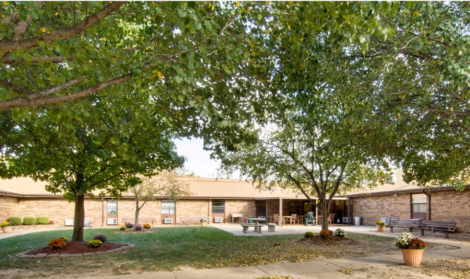 Courtyard of a single-story brick nursing center with large shade trees, picnic tables, benches, and potted flowers.