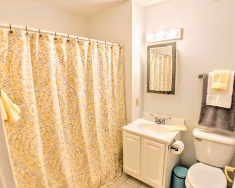 A bathroom with a patterned yellow and white shower curtain, a white sink vanity with a mirror above it, a toilet with folded towels on the towel rack above, and a small blue trash bin on the floor.