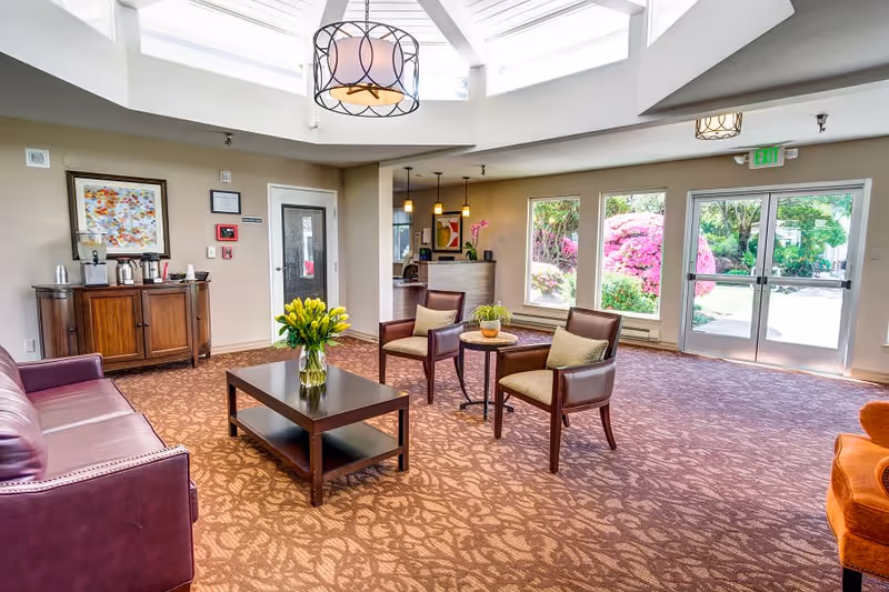 A bright and spacious senior living facility common area with a patterned carpet, a purple leather sofa, two armchairs with cushions around a small round table, and a rectangular coffee table with a vase of yellow flowers. There is a wooden cabinet with coffee supplies against the wall, large windows showing colorful flowering bushes outside, and a glass door leading outdoors. The ceiling features a skylight and a decorative hanging light fixture.