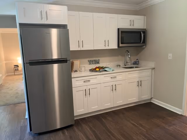 A modern kitchen area with white cabinets, a stainless steel refrigerator, a built-in microwave above a sink, and a countertop with a fruit bowl, a cup, and a decorative 'YUM' sign. The floor is dark wood, and there is an adjacent carpeted room visible through an open doorway.