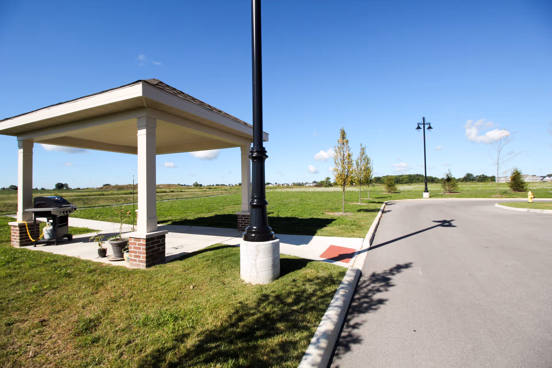 A small covered pavilion with a grill sits beside a road and lampposts in a grassy outdoor area under a clear blue sky.