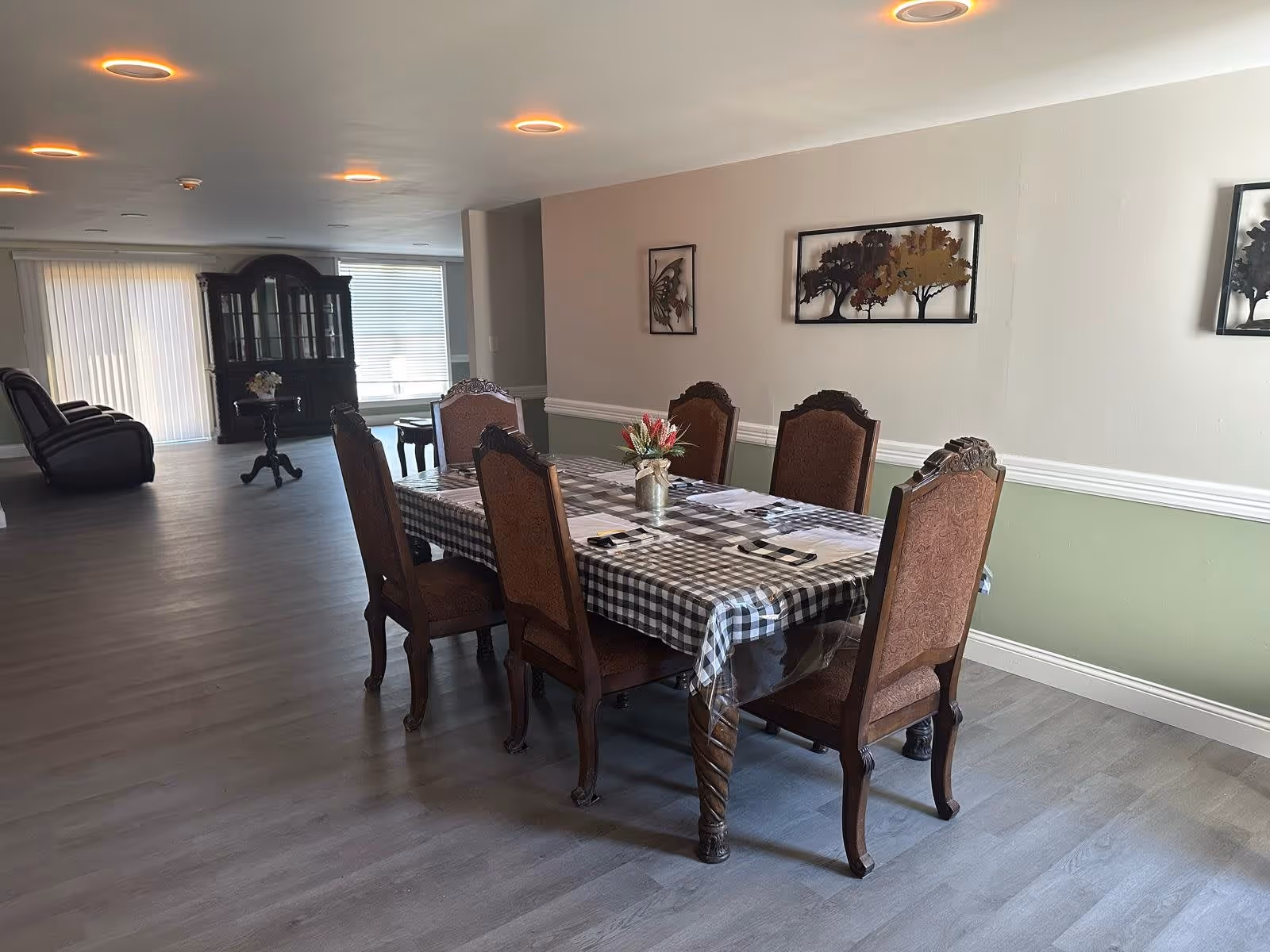 A dining area in a senior living facility with a rectangular table covered with a checkered tablecloth and six wooden chairs with upholstered seats and backs. A small flower arrangement is placed in the center of the table. The room has light-colored walls with decorative wall art featuring trees and a butterfly. In the background, there is a dark wooden cabinet and two recliner chairs near large windows with vertical blinds. The floor is covered with light gray wood-like flooring.