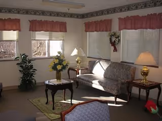 A cozy living room with a floral-patterned sofa, two wooden side tables with brass lamps, a small coffee table with a vase of yellow flowers, and windows with red valances letting in natural light. There is also a green plant in the corner and a decorative wall hanging.