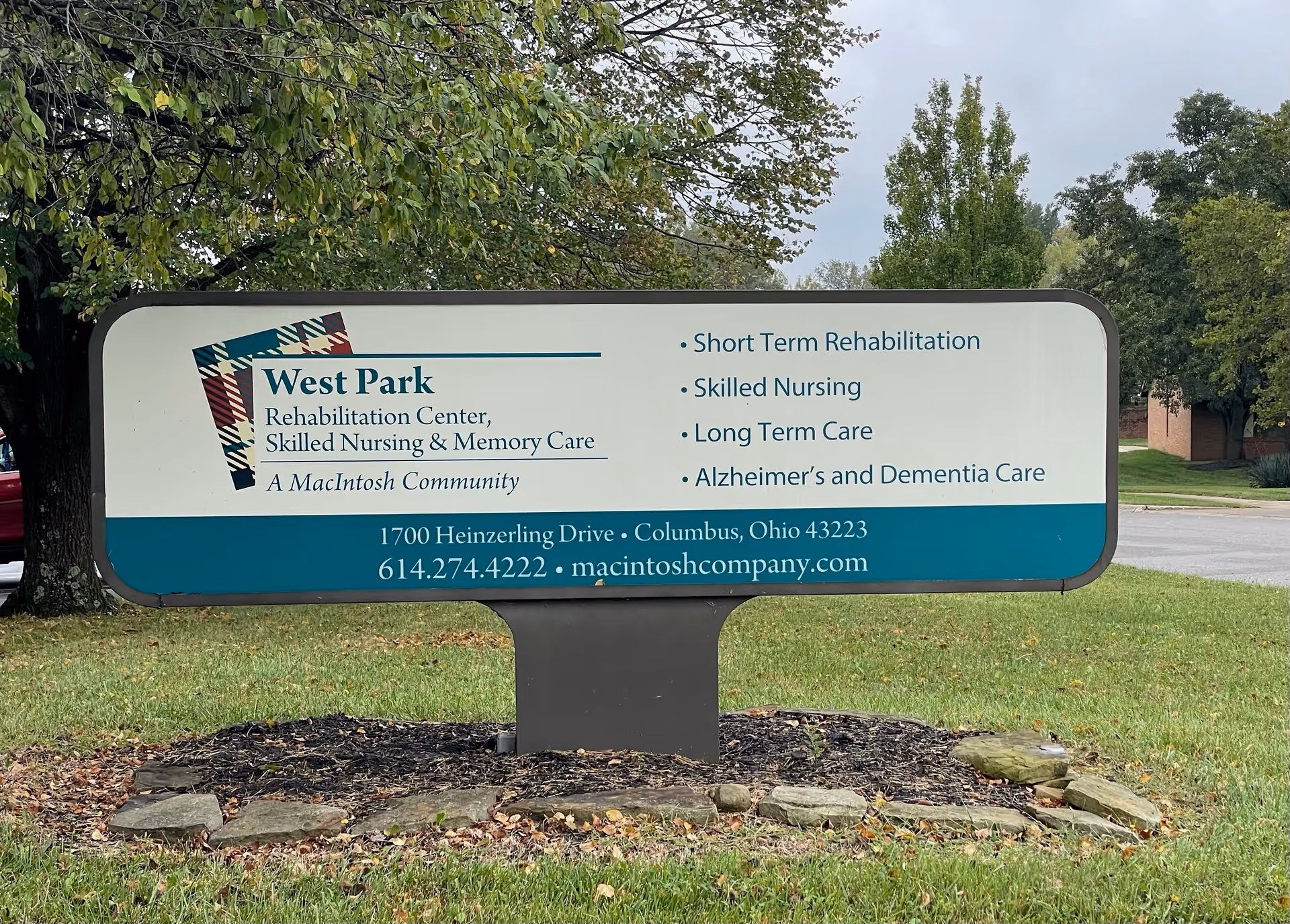 Outdoor sign for West Park Rehabilitation Center, Skilled Nursing & Memory Care, listing services including short term rehabilitation, skilled nursing, long term care, and Alzheimer's and dementia care. The sign includes the address 1700 Heinzerling Drive, Columbus, Ohio 43223, phone number 614.274.4222, and website macintoshcompany.com. The sign is situated on a grassy area with trees and a parking lot in the background.