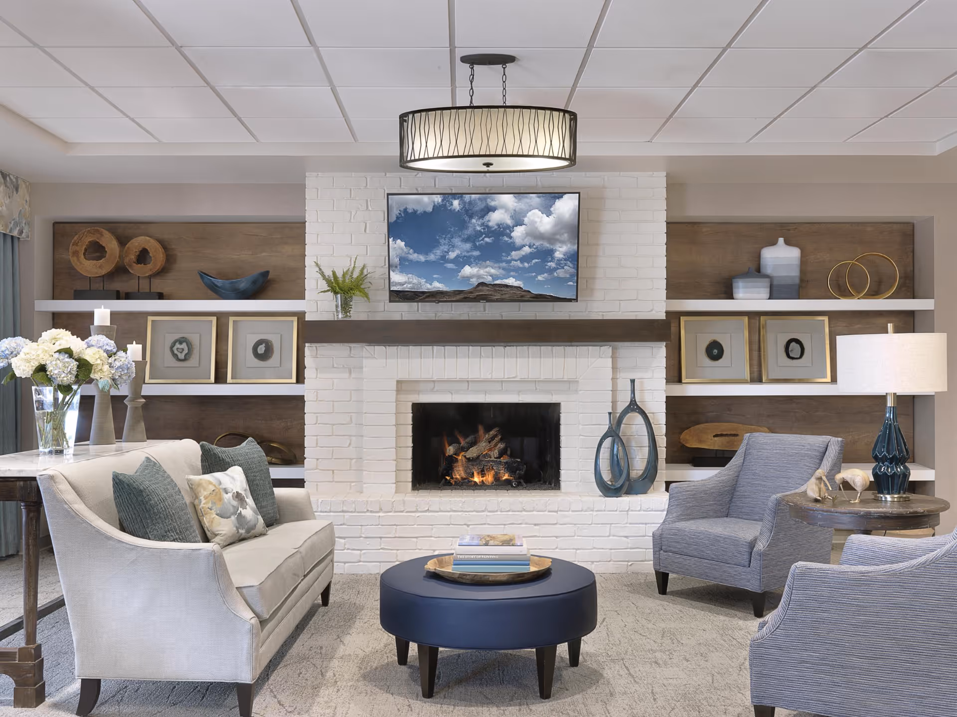 A cozy living room with a white brick fireplace in the center, a flat-screen TV mounted above it displaying a cloudy sky, and decorative shelves on either side holding framed art and sculptures. The room features a beige sofa with cushions, two gray armchairs, a round blue ottoman coffee table with books on it, a side table with a lamp, and a vase of flowers on a wooden table to the left.