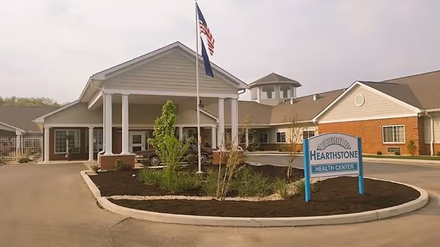 Front exterior view of Hearthstone Health Center building with a covered entrance supported by white columns, an American flag and another flag on flagpoles, landscaped area with plants and mulch, and a sign reading 'Hearthstone Health Center'.