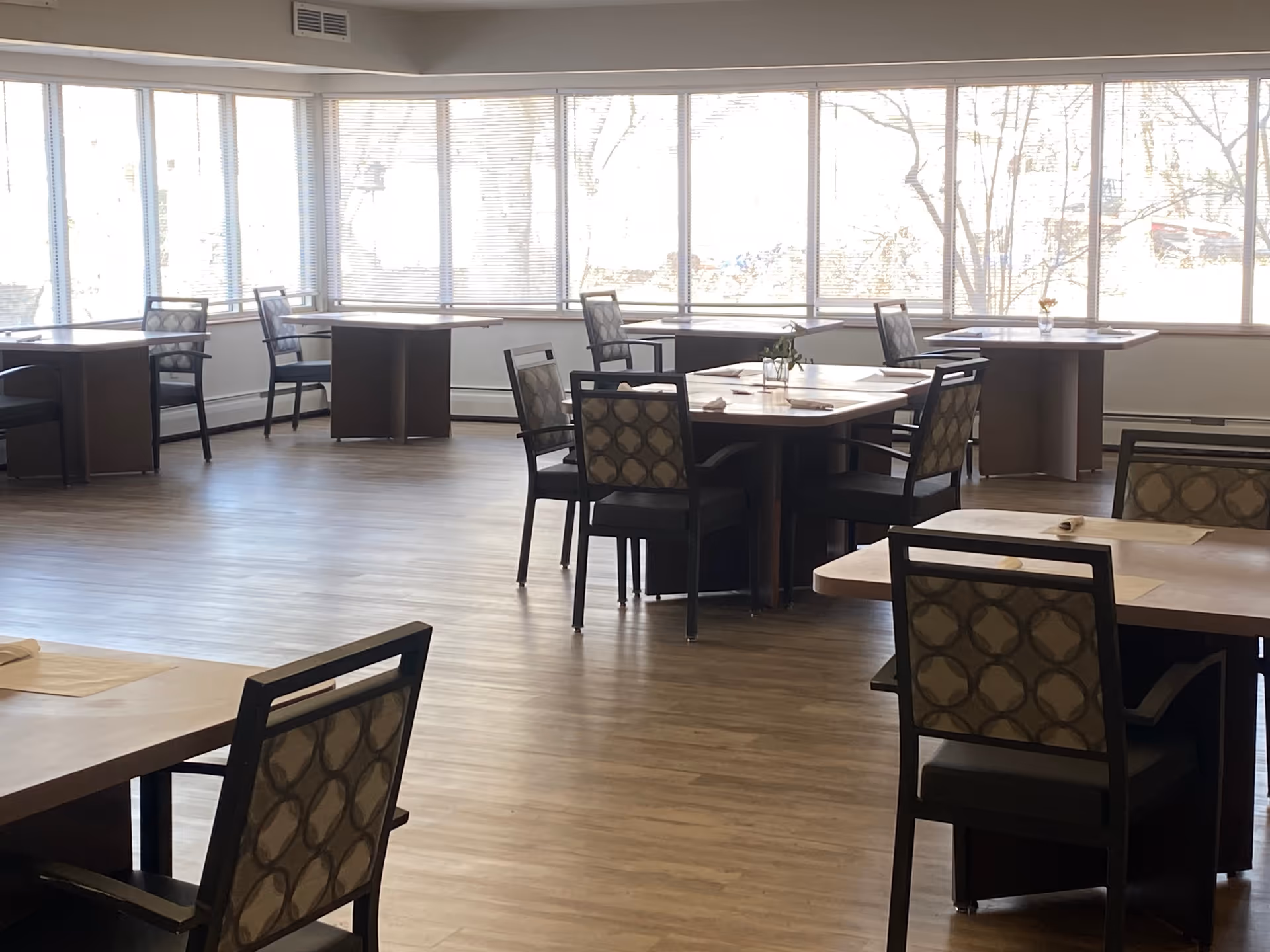 A bright dining room with several square wooden tables and cushioned chairs arranged neatly. Large windows with blinds cover the walls, letting in natural light and showing a view of trees outside. The floor is wooden, and some tables have small vases with flowers.