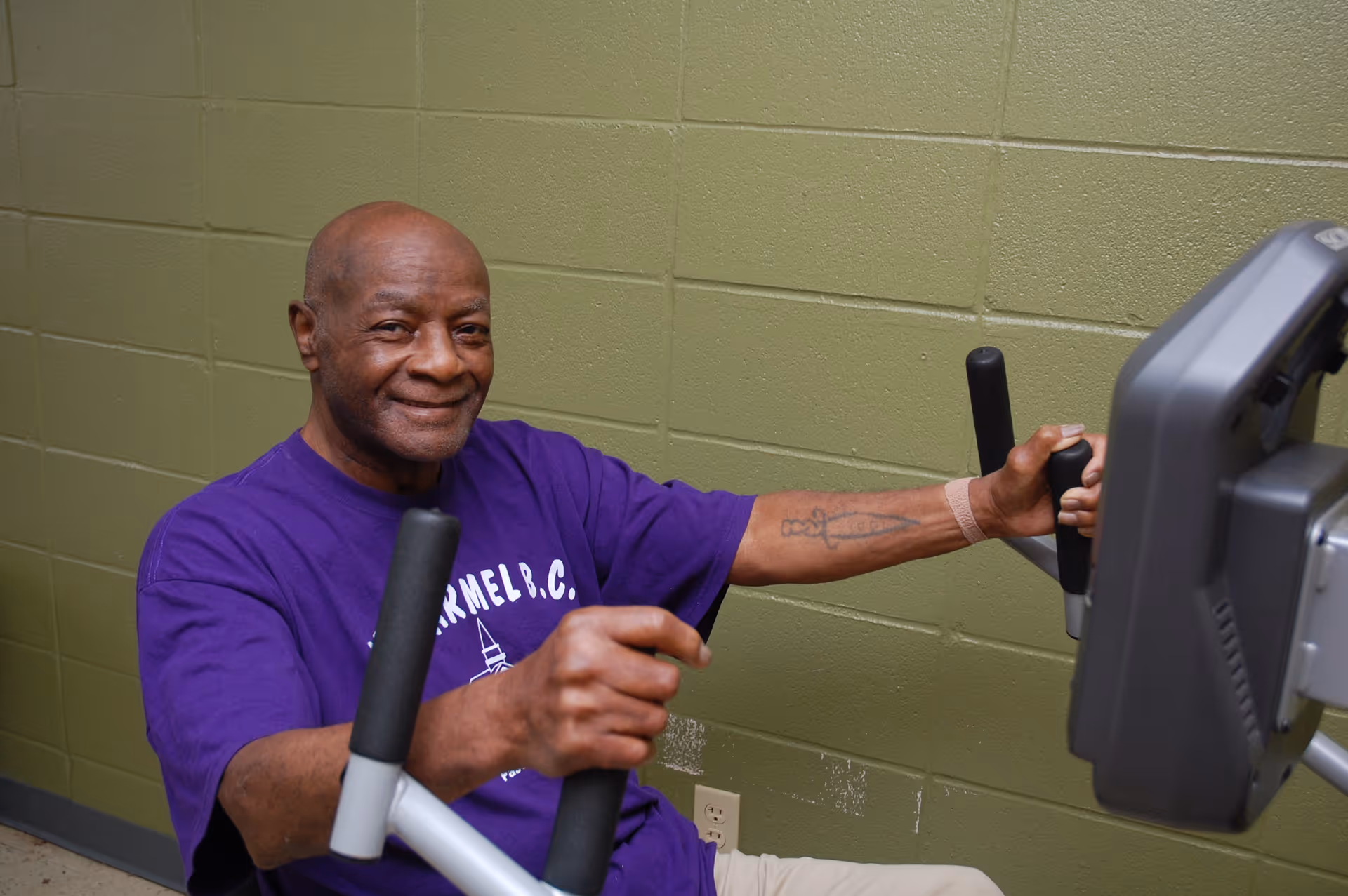 An elderly man wearing a purple shirt is exercising on a seated chest press machine against a green cinder block wall, smiling at the camera.
