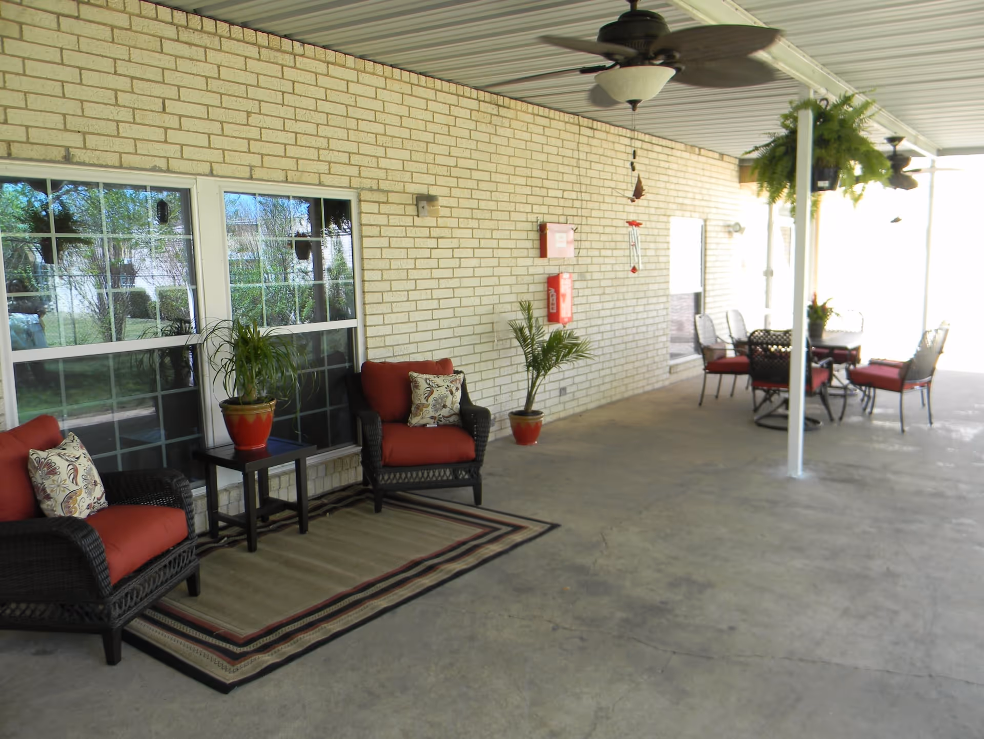 Covered outdoor patio area with brick walls, ceiling fans, and seating arrangements including cushioned chairs and a table. There are potted plants and a fire extinguisher mounted on the wall.