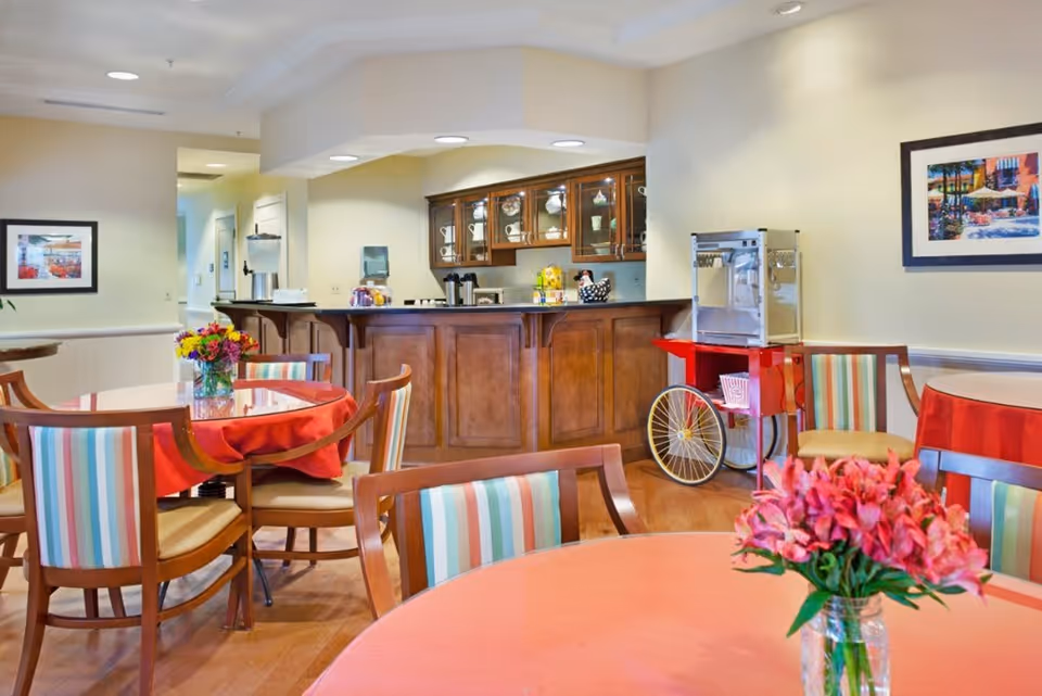 A cozy dining area in a senior living facility with round tables covered in red tablecloths, wooden chairs with striped cushions, a wooden counter with glass cabinets displaying dishes, a popcorn machine on a red cart, and framed artwork on the walls.