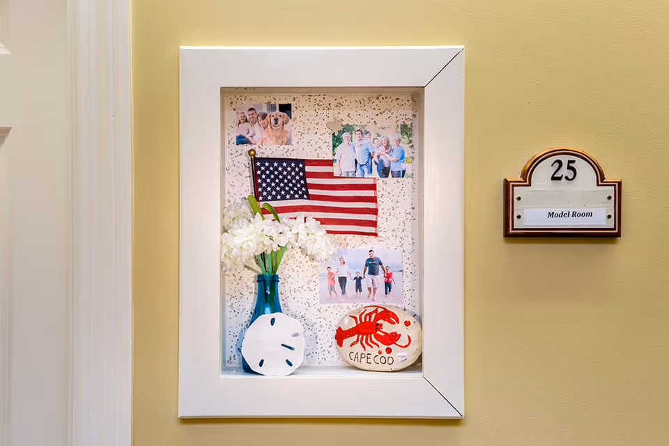 Decorative wall niche in a hallway displaying an American flag, family photos, a vase of white flowers and a Cape Cod painted rock next to a room plaque numbered 25 labeled "Model Room".