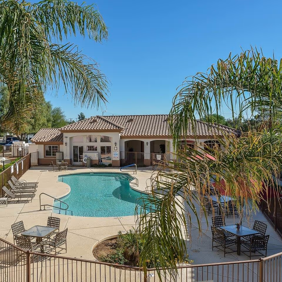 Outdoor swimming pool area at Ventana Winds Retirement Home with lounge chairs, tables with chairs, palm trees, and a building with a tiled roof in the background under a clear blue sky.