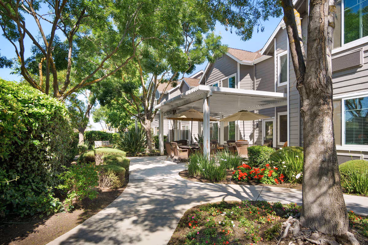 Outdoor patio area at The Grove Senior Living with a paved walkway, surrounded by trees, bushes, and colorful flowers. There are several wicker chairs and tables under a white pergola attached to a gray building with multiple windows.