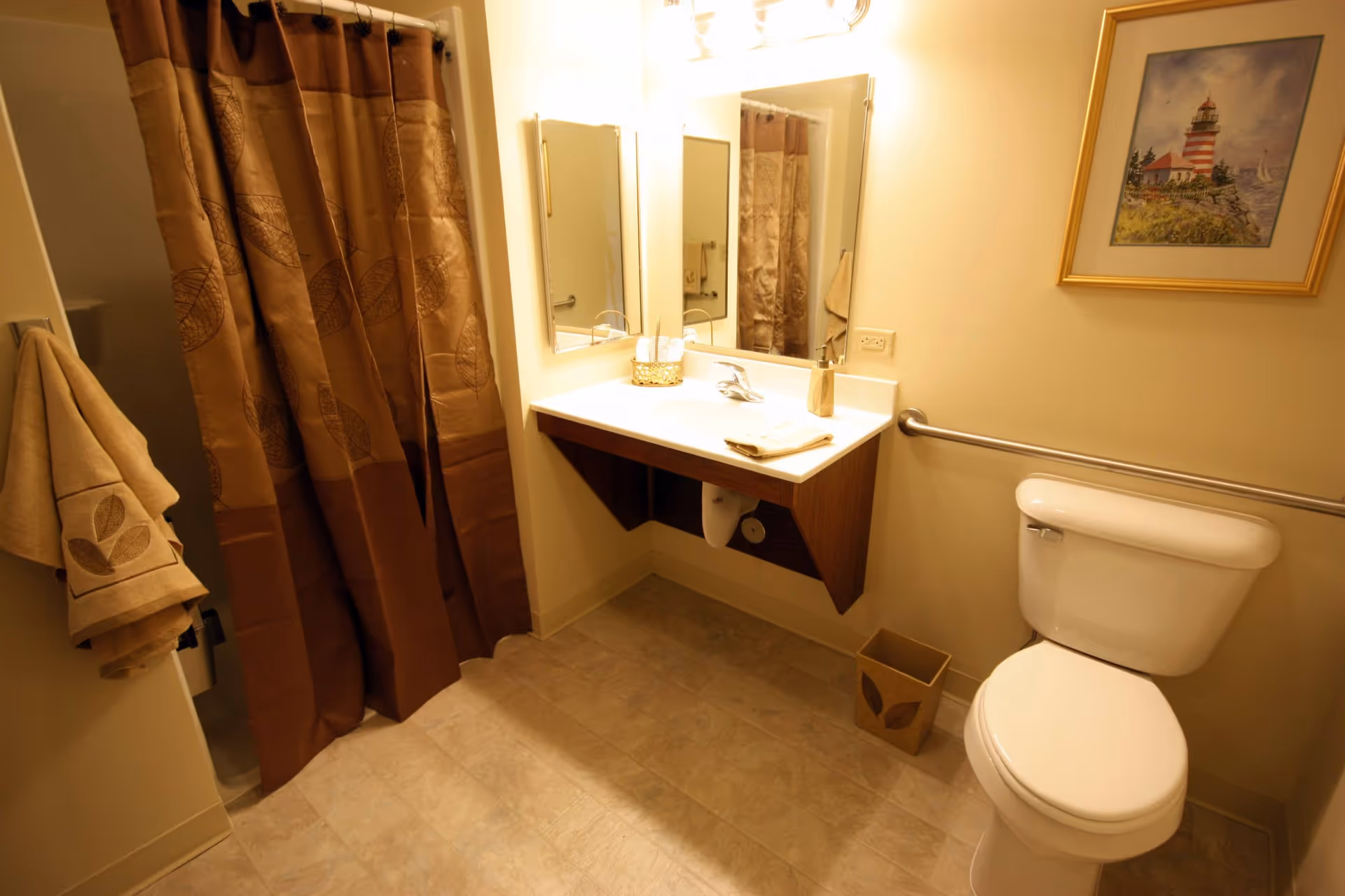 A bathroom with a white toilet, a wall-mounted sink with a mirror above it, a brown shower curtain with leaf patterns, beige towels hanging on a rack, a small trash bin, and a framed picture of a lighthouse on the wall.