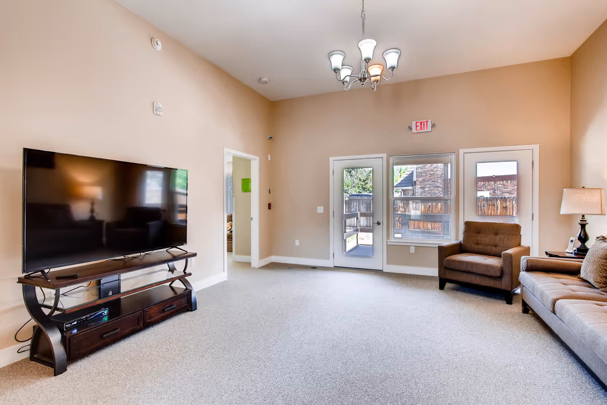 Bright common living room with a large TV on a stand, sofa and armchair, carpeted floor, and doors/windows leading outside.