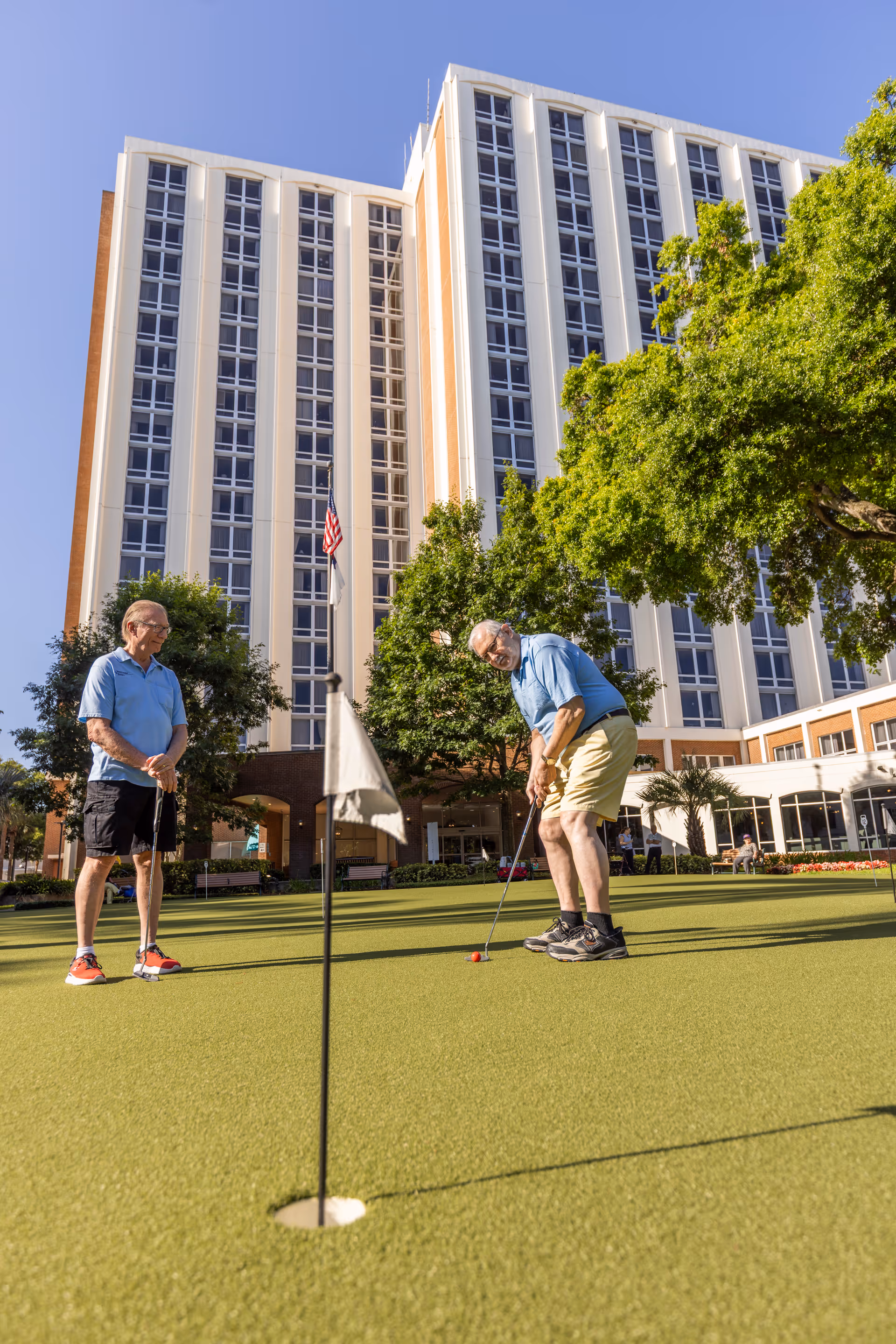 Two older men putting on a green in front of a tall multi-story residential building.