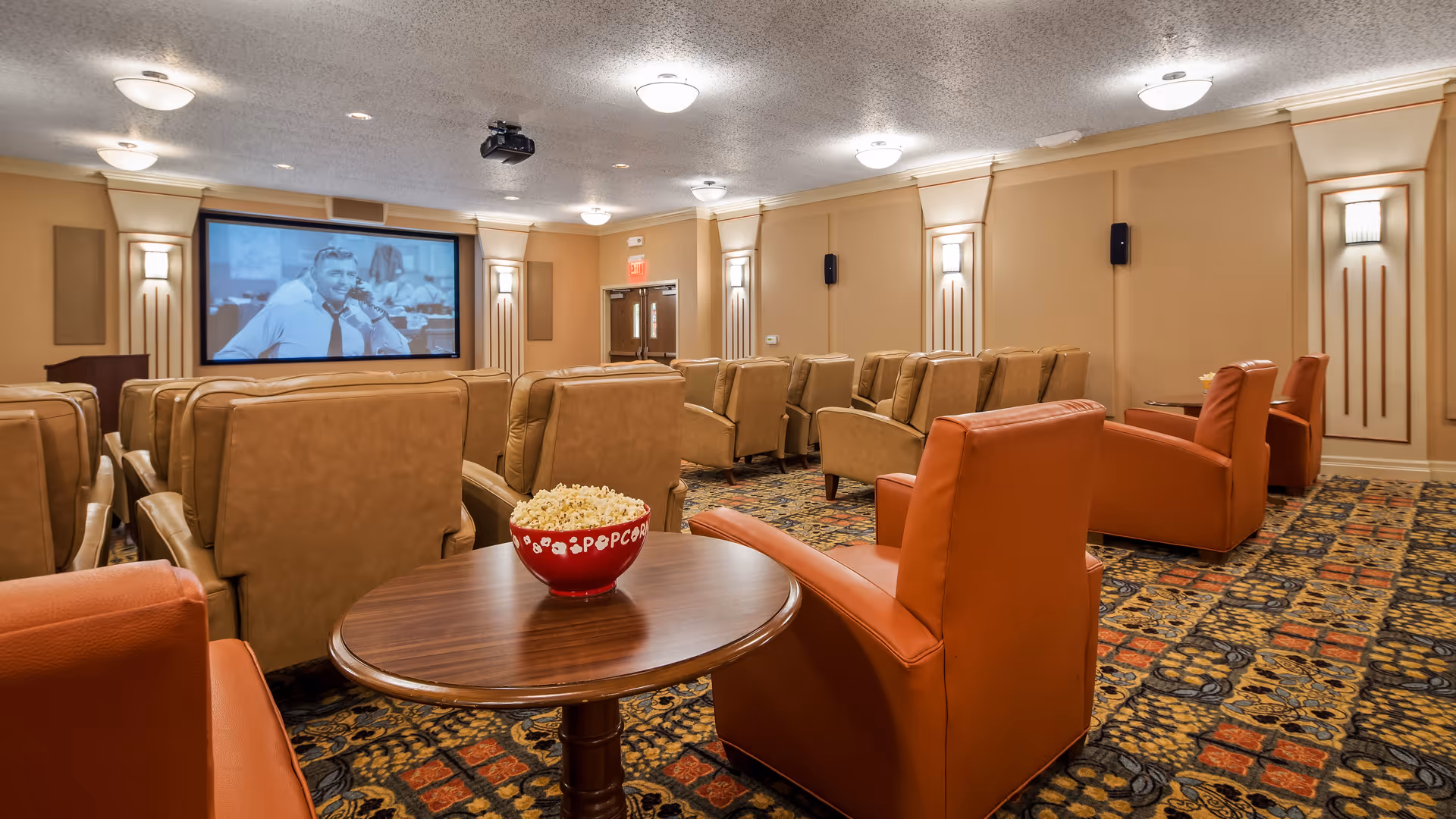 A cozy movie theater room with multiple rows of comfortable tan and orange armchairs facing a large screen showing a black and white movie. A round wooden table in the foreground holds a red bowl filled with popcorn. The room has warm lighting and patterned carpet.