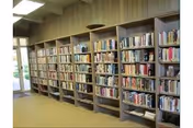 Interior view of a library or reading room with multiple wooden bookshelves filled with books along the wall. There is a large window or glass door on the left side allowing natural light to enter the room.