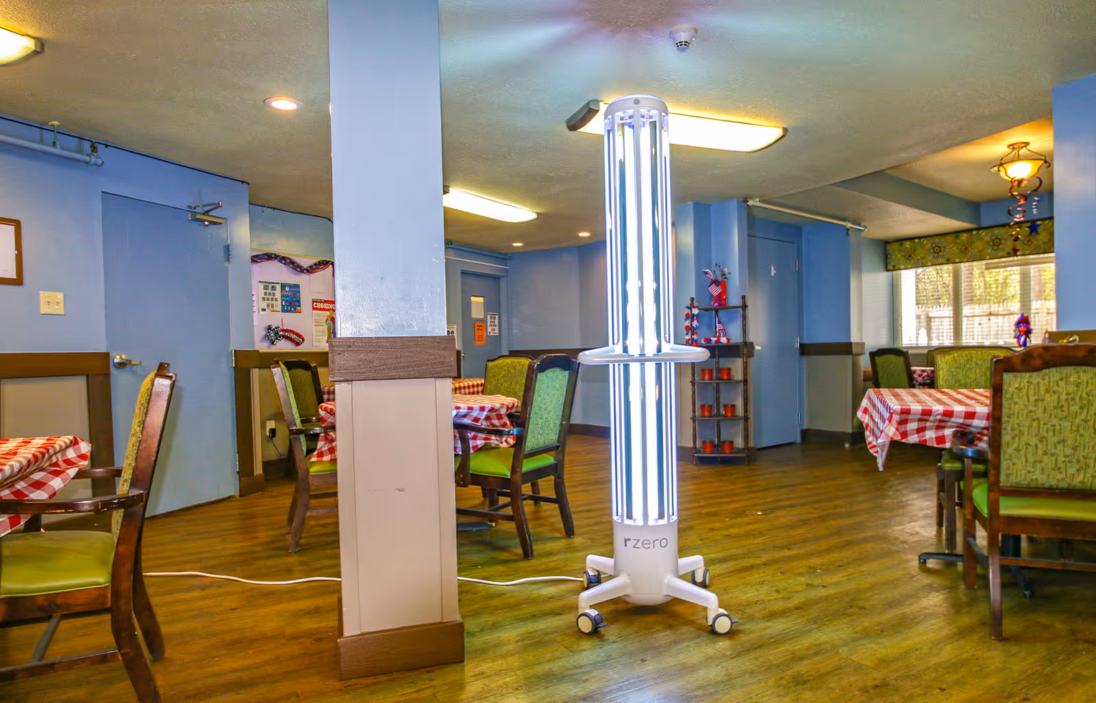 Interior of a dining room with several tables covered in red and white checkered tablecloths and green cushioned chairs. A tall, white UV disinfection lamp labeled 'rzero' is positioned in the center of the room. The room has blue walls, wooden flooring, and windows with patterned valances letting in natural light.