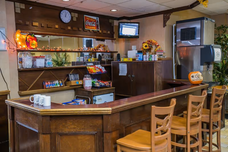 Interior view of a small snack bar or refreshment area with a wooden counter and three wooden chairs. Behind the counter are shelves stocked with snacks, a small refrigerator, a coffee machine, and a television mounted on the wall. The area is decorated with a pumpkin, a fall-themed wreath, and string lights.