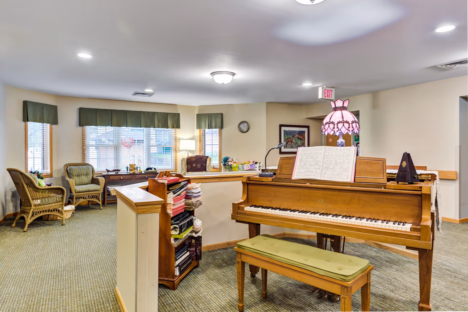 Bright common room featuring an upright piano and bench, wicker seating, and a windowed reading nook.