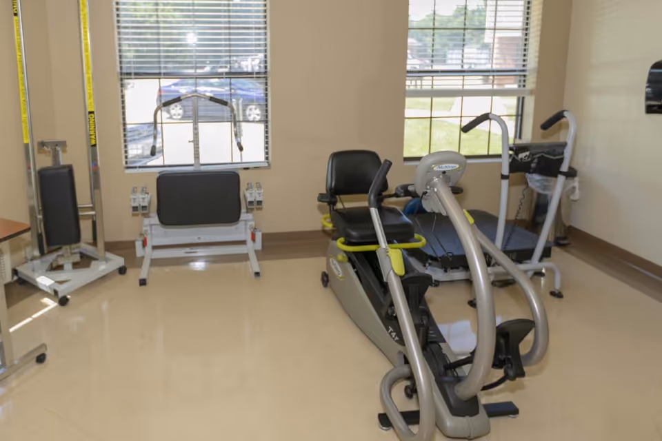 A small exercise room with various fitness equipment including a recumbent exercise bike, a treadmill, and a weight machine. The room has beige walls, two windows with blinds, and a shiny tiled floor.