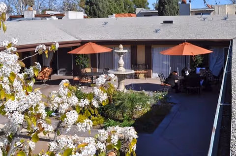 Outdoor courtyard area with a central stone fountain surrounded by greenery and flowering trees. There are two orange patio umbrellas shading seating areas where people are sitting and conversing. The courtyard is enclosed by a building with large windows and sliding doors.
