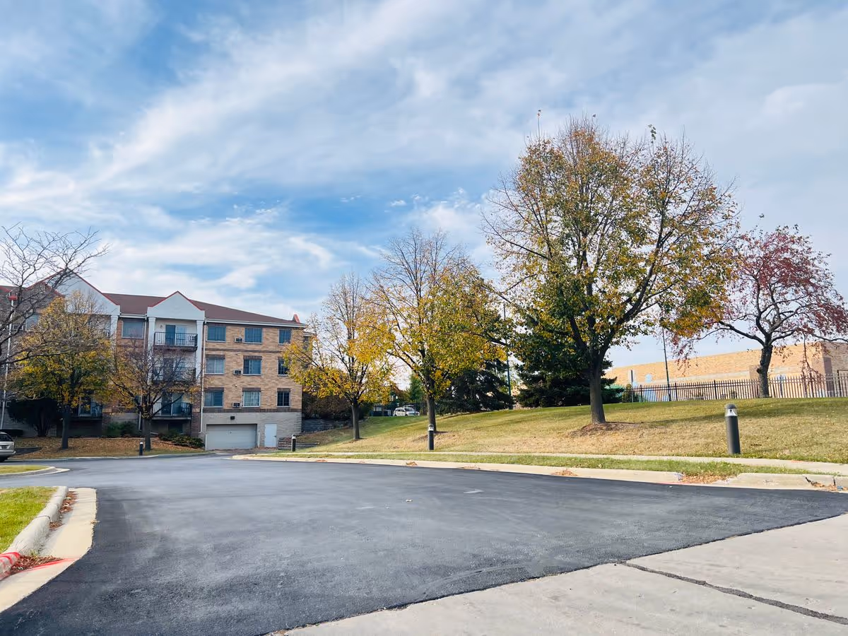 Front exterior of a multi-story residential building with a paved driveway, lawn and trees under a blue sky.