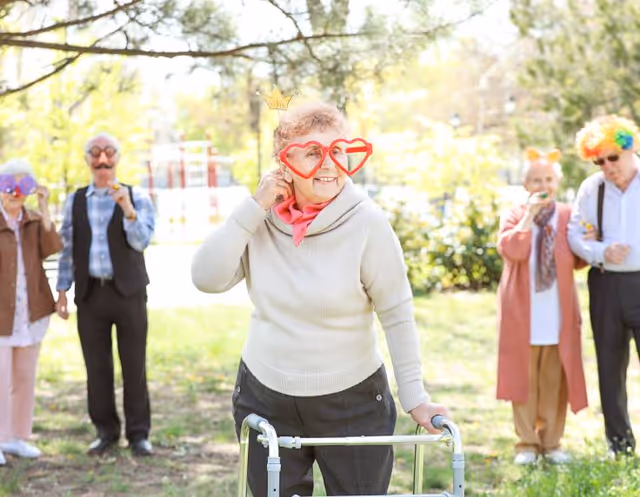 A group of elderly people outdoors in a park-like setting wearing playful costume accessories such as oversized glasses, a crown, and a colorful wig. One woman in the foreground uses a walker and wears large red heart-shaped glasses and a small crown, smiling and looking to the side. The others stand in the background also wearing fun accessories.
