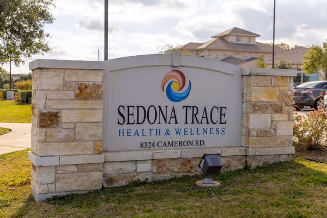 Stone sign at the entrance of Sedona Trace Health & Wellness facility with the address 8324 Cameron Rd, surrounded by grass and a building in the background under a partly cloudy sky.