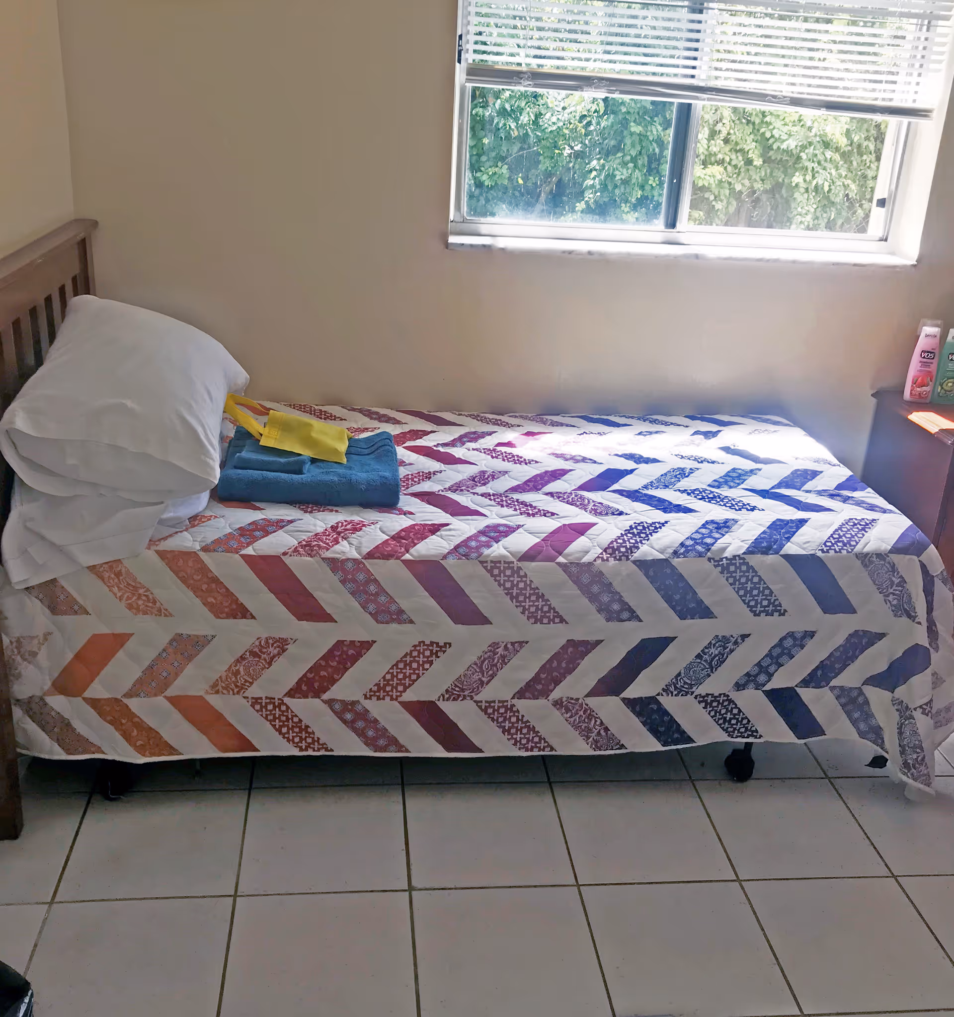 A single bed with a colorful patterned quilt and two white pillows stacked at the head. On the bed are folded blue towels and a yellow item. The bed is positioned against a beige wall under a window with white blinds partially open, showing green foliage outside. To the right of the bed is a small wooden nightstand with various personal care items on top. The floor is tiled with white tiles.