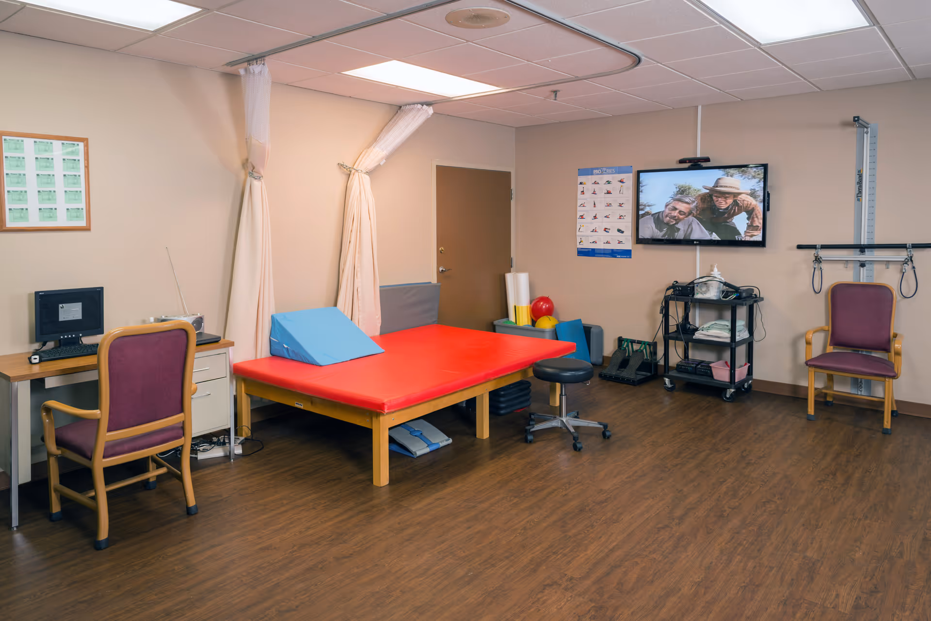A therapy room with a red padded treatment table, a blue wedge cushion, two chairs, a computer desk with a monitor, a TV mounted on the wall displaying two people, exercise equipment, and a poster with exercise illustrations. The room has wood flooring and beige walls.