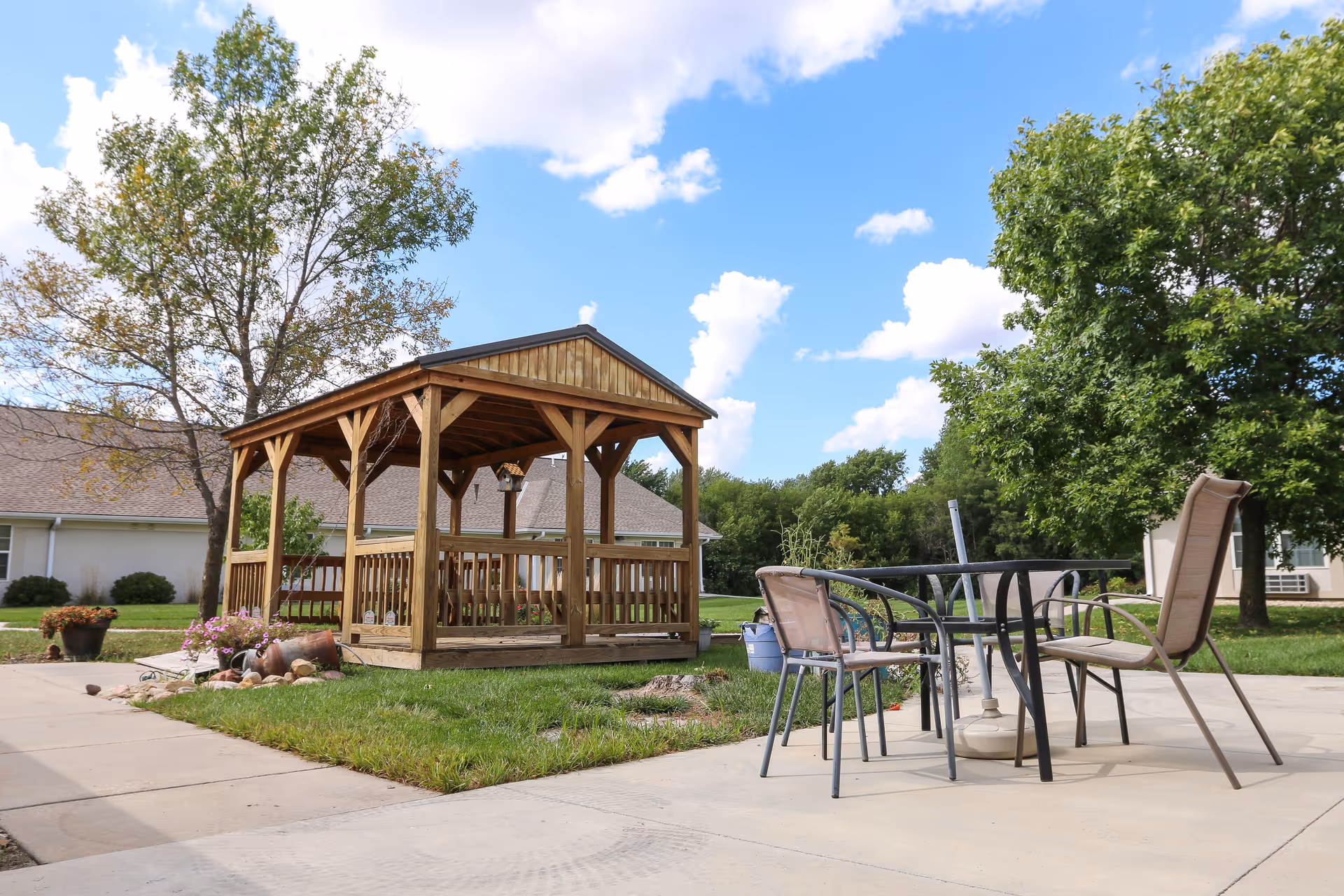 Outdoor patio area at Vintage Park at Hiawatha featuring a wooden gazebo with a bench swing inside, surrounded by green grass and trees. There is a round metal table with four chairs on a concrete patio under a partly cloudy blue sky.