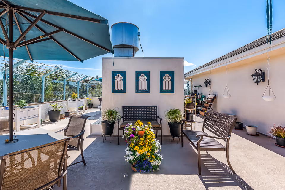 Outdoor patio area with metal benches, a table with chairs, a large green umbrella, potted plants, and decorative wall art under a clear blue sky.