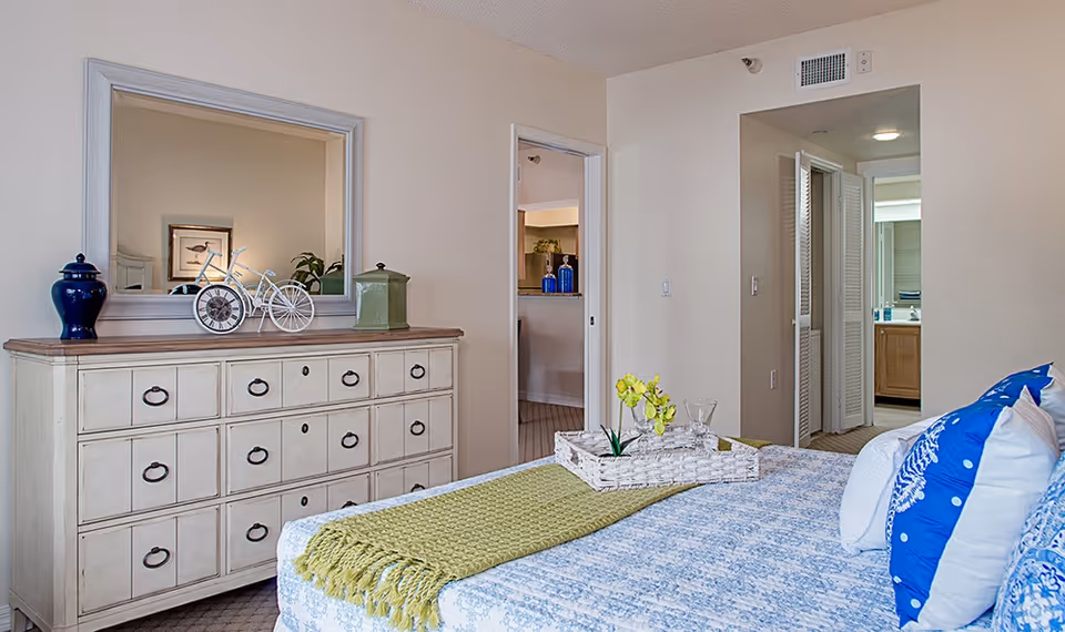 A bedroom with a bed covered in a blue and white patterned bedspread and blue and white pillows. A green throw blanket is folded at the foot of the bed with a white tray holding a small plant and glassware. Across from the bed is a cream-colored dresser with multiple drawers, decorated with a blue vase, a decorative bicycle, and a green container. A large mirror hangs above the dresser. The room has beige walls and carpeted floors. Two doorways lead to other rooms, one showing a glimpse of a kitchen area and the other a bathroom with a sink and mirror.