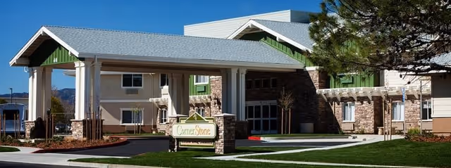 Exterior view of Cornerstone Assisted Living facility showing the main entrance with a covered drop-off area, stone and siding facade, and a sign with the facility name in front on a well-maintained lawn.
