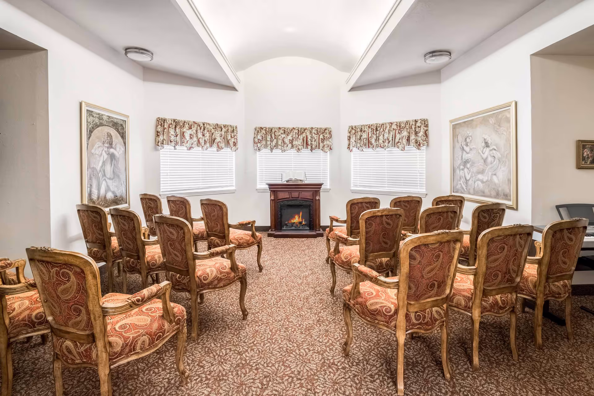 A small meeting or gathering room with two rows of ornate wooden chairs upholstered in red patterned fabric facing a small electric fireplace. The room has three windows with white blinds and floral valances. Two framed paintings hang on the walls, and the carpet has a brown and beige pattern.