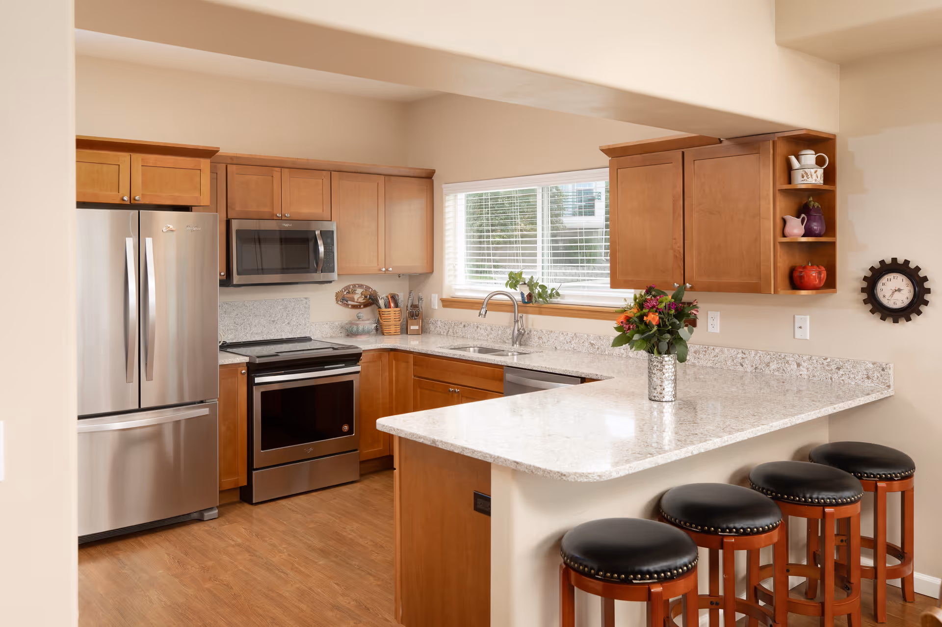 A modern kitchen with wooden cabinets, stainless steel refrigerator, oven, and microwave. There is a large window above the sink letting in natural light. A granite countertop extends into a breakfast bar with four wooden stools with black cushioned seats. A vase with colorful flowers is placed on the countertop. A decorative clock is mounted on the wall.