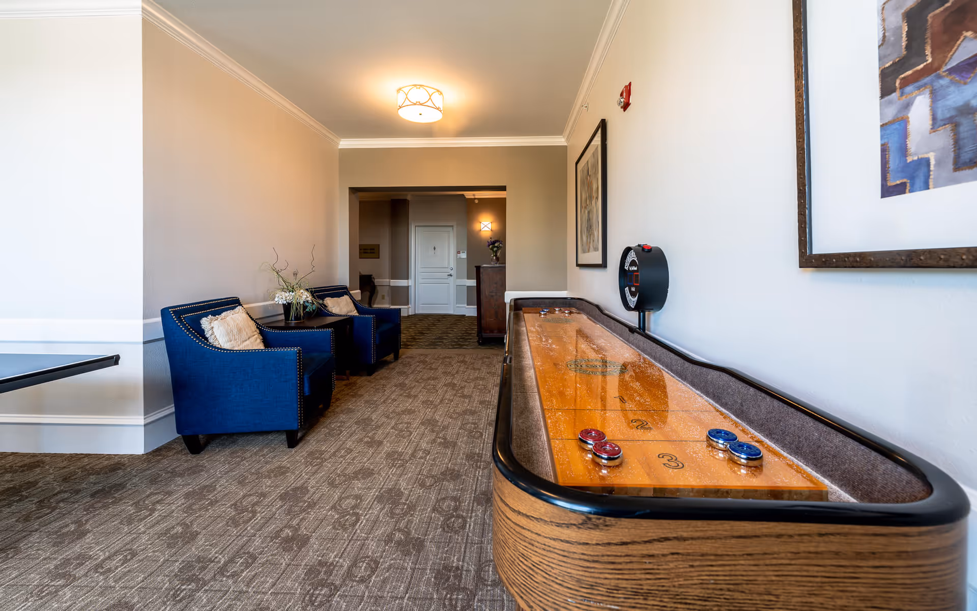 Interior view of a senior living facility game room featuring a shuffleboard table on the right, two blue armchairs with beige pillows and a small table with a flower arrangement on the left, beige walls, carpeted floor, and framed artwork on the wall.