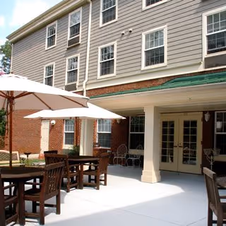 Outdoor patio area at Historic Roswell Place featuring wooden tables and chairs with large white umbrellas, adjacent to a multi-story building with beige siding and multiple windows.