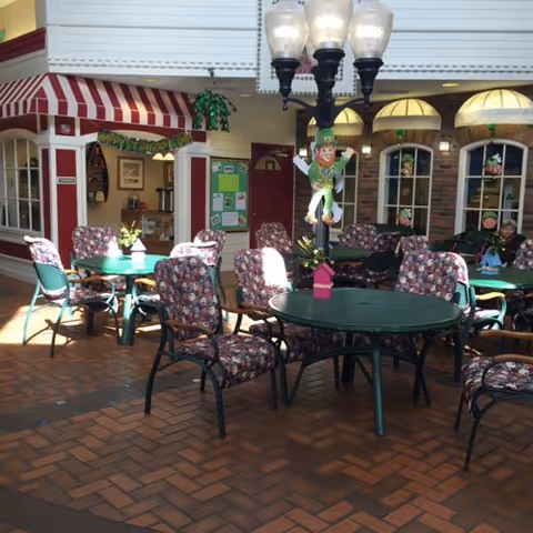 Indoor seating area with round green tables and floral cushioned chairs arranged on a brick floor. A decorative lamppost with three lights and a hanging leprechaun figure is in the center. The background shows a small storefront with a red and white striped awning and windows with stained glass decorations.