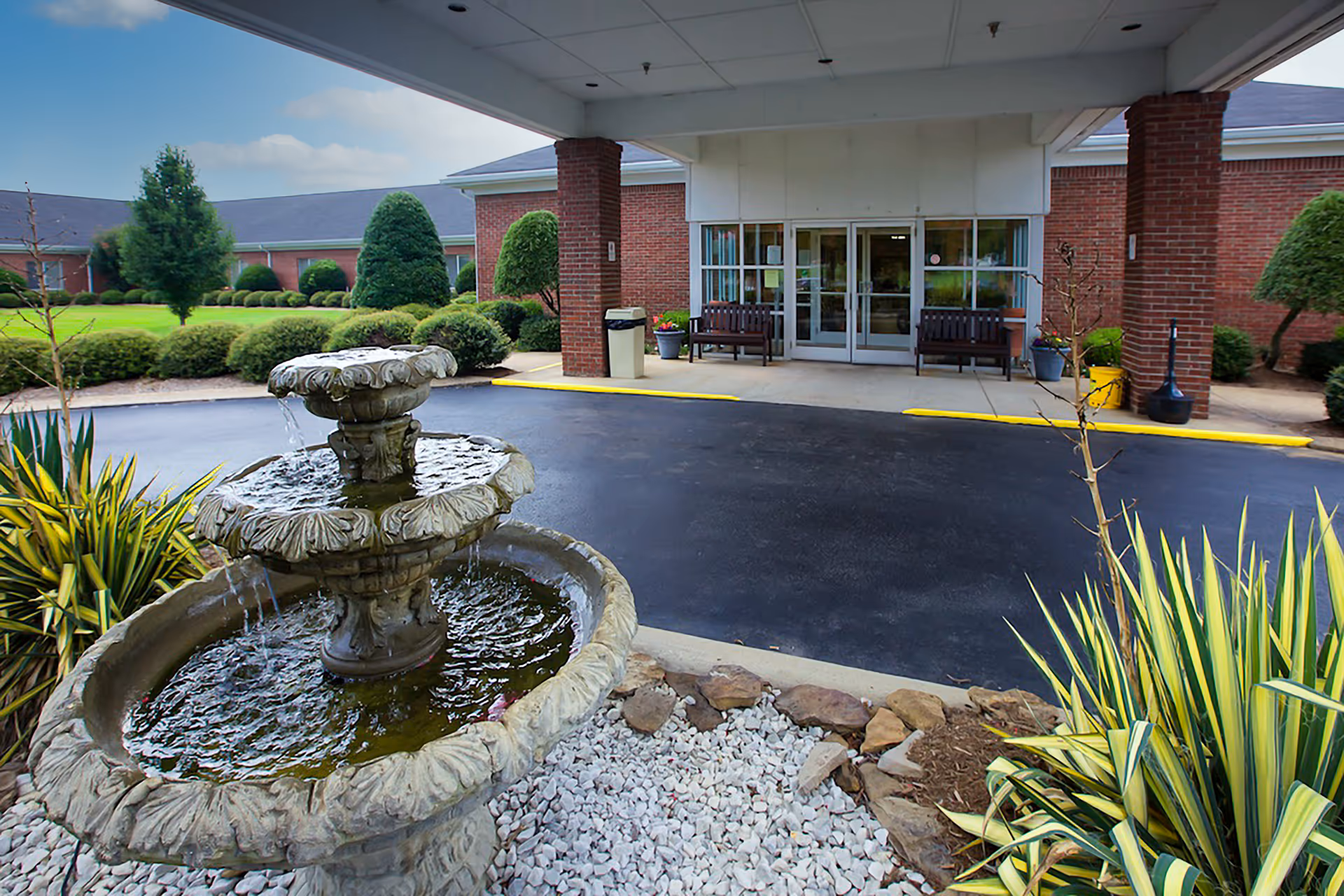 Outdoor view of the entrance to Cherrywood Post Acute facility featuring a two-tier stone water fountain in the foreground, surrounded by landscaping with green shrubs and plants. The building has a covered drop-off area with benches near the glass doors.