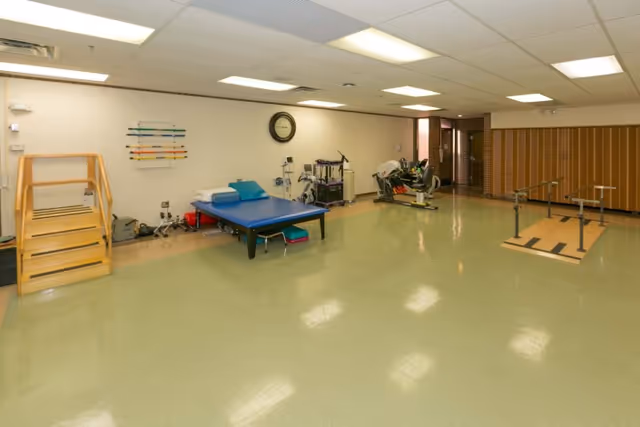 A spacious rehabilitation room with a green floor and white ceiling lights. The room contains physical therapy equipment including a wooden staircase, a blue therapy table, exercise machines, parallel bars for walking practice, and various therapy tools mounted on the wall. A clock is mounted on the wall above the therapy table.
