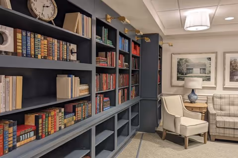 Interior view of a cozy reading or sitting area with built-in dark gray bookshelves filled with books and a clock on the top shelf. There is a beige armchair and a plaid sofa next to a wooden side table with a blue lamp. Two framed pictures hang on the wall above the sofa, and a ceiling light fixture is visible.