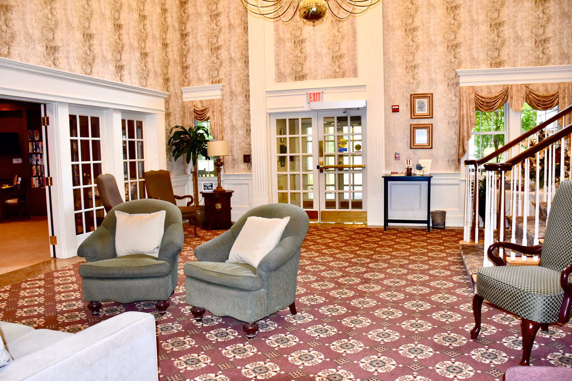 A spacious and elegant common area with patterned carpet, two green armchairs with white pillows, a wooden side table with a lamp, and a staircase with white railings. There are large windows with drapes, double glass doors with an exit sign above, and framed pictures on the wall.