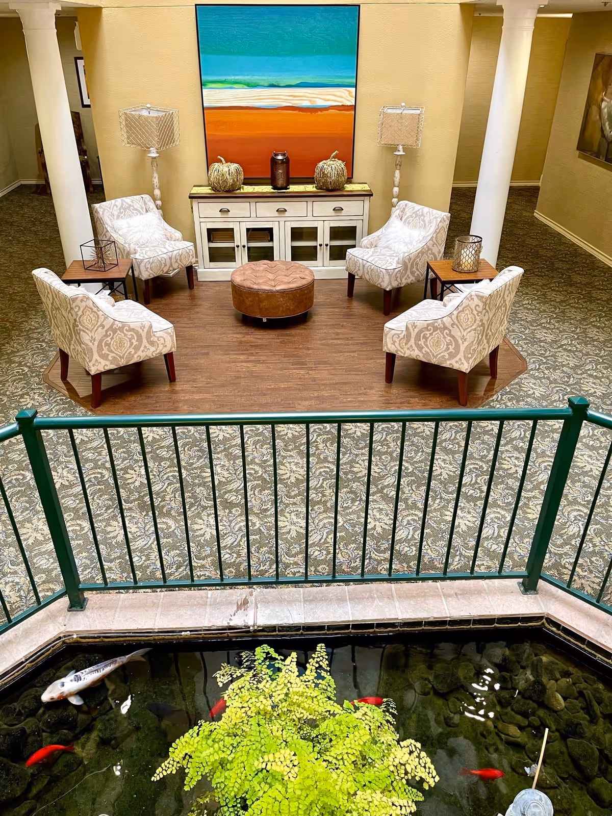 Indoor seating area with four patterned armchairs arranged around a round brown ottoman on a wooden floor section. Behind the chairs is a white cabinet with decorative pumpkins and a lantern on top, flanked by two floor lamps with patterned shades. A large colorful abstract painting hangs on the wall above the cabinet. In the foreground, a green metal railing surrounds a small indoor pond with fish and a plant.